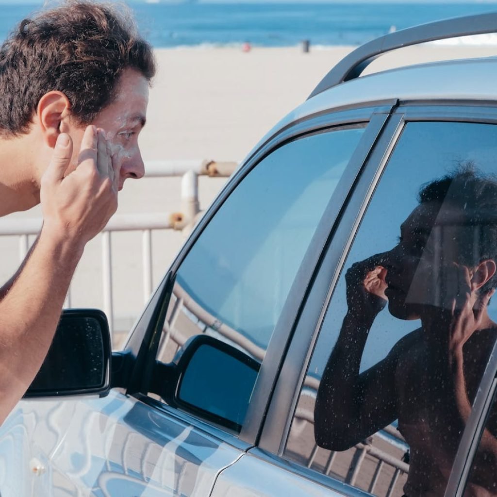 A man applies sunscreen using his car window reflection at a sunny beach.