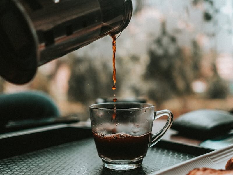 Aromatic coffee being poured into a glass cup beside a plate of biscotti on a tray.