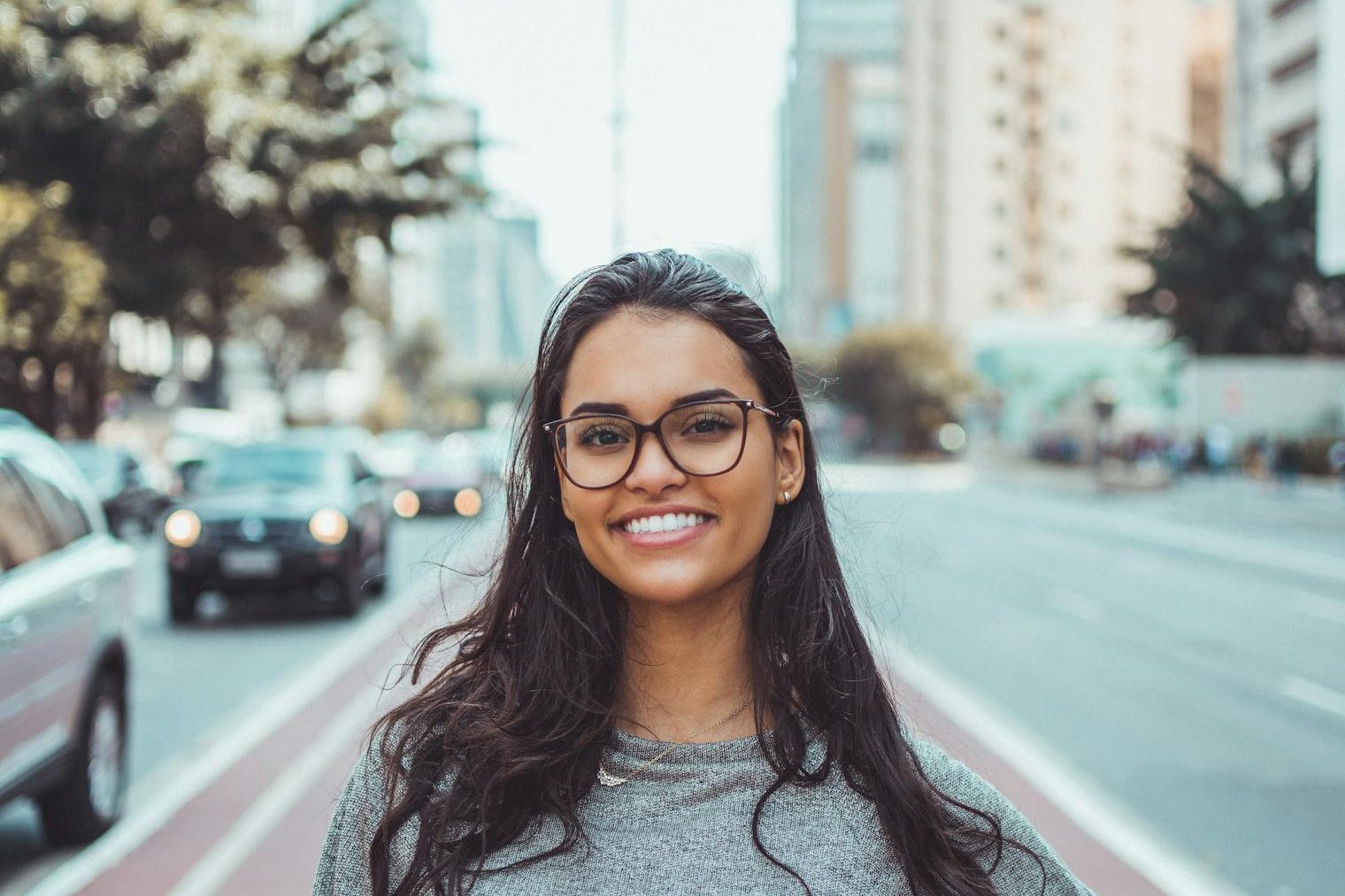 A young woman with glasses smiling on a city street, embracing urban lifestyle.